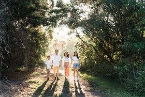 Family of four walking hand-in-hand on a sunlit forest path surrounded by lush green trees.