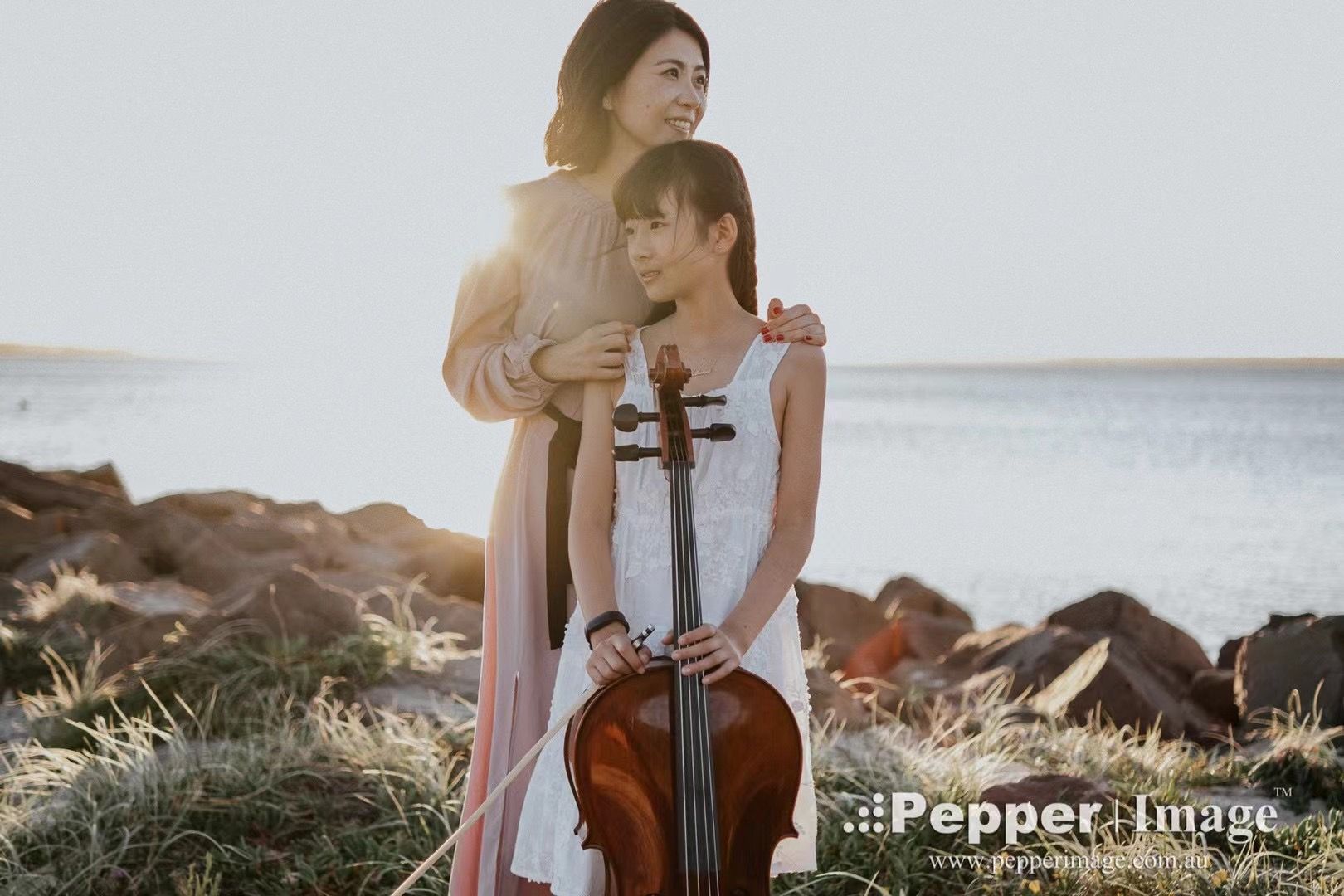 Two women pose outdoors near water, one holding a violin, in warm golden sunlight among rocks and grass.