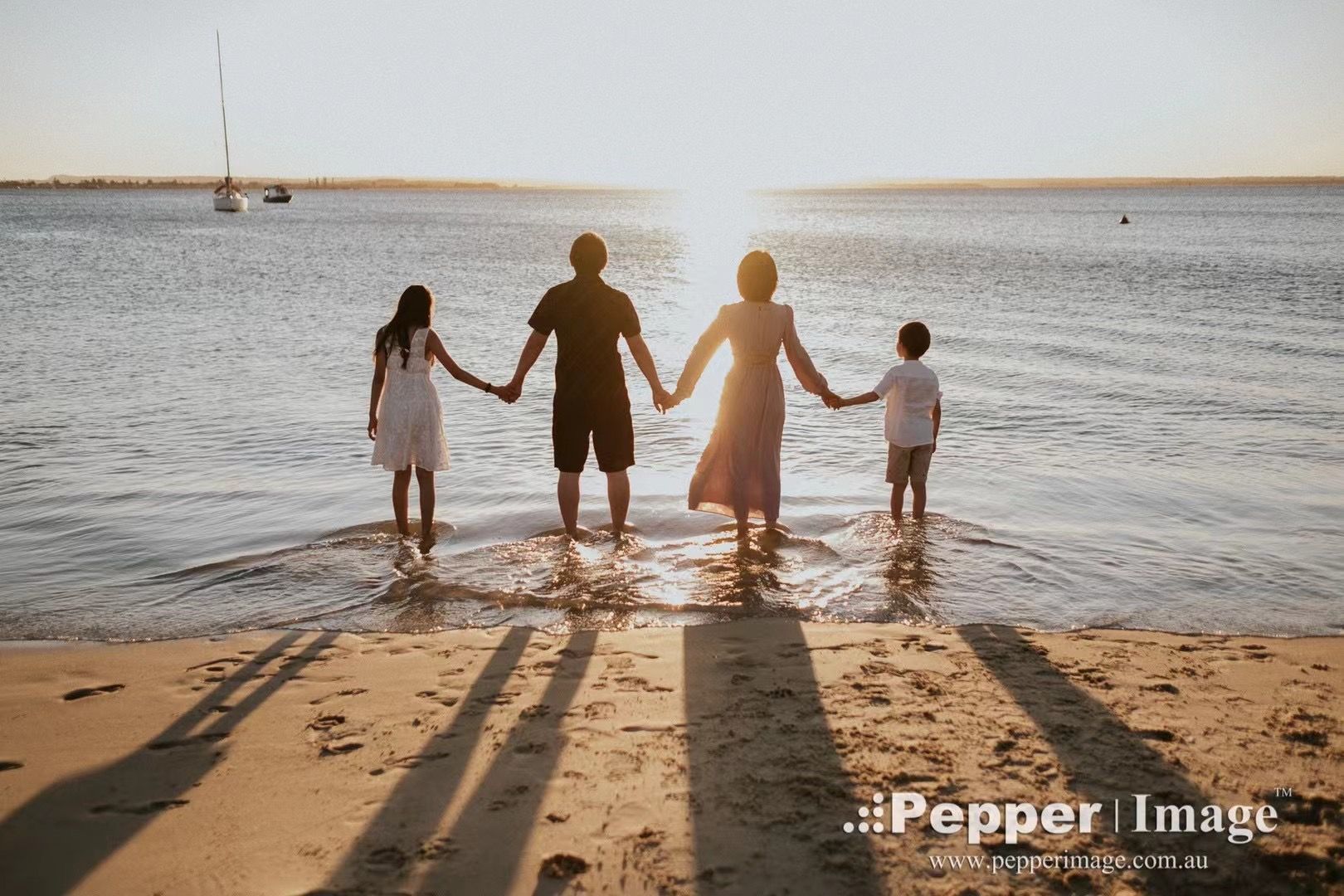 Family of four holding hands at sunset on a beach, silhouetted against golden light reflecting on calm water.