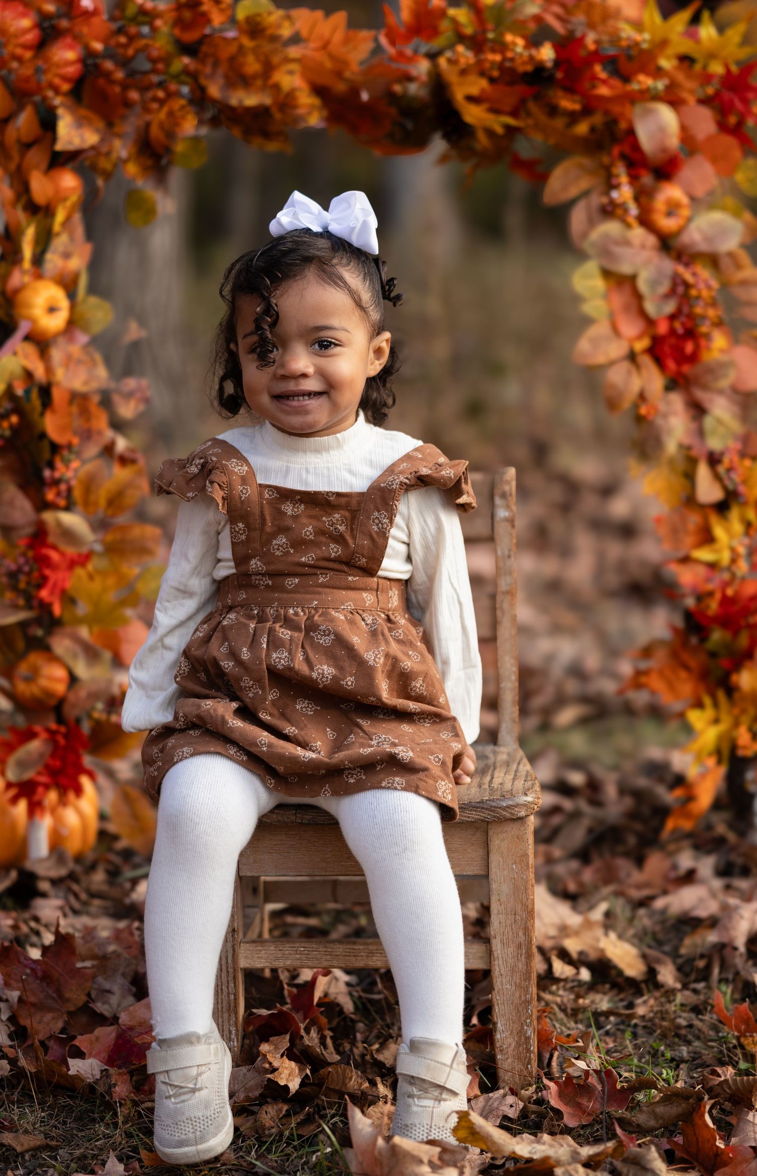 A smiling toddler girl in a brown dress sits on a chair under a fall foliage arch for autumn portraits.