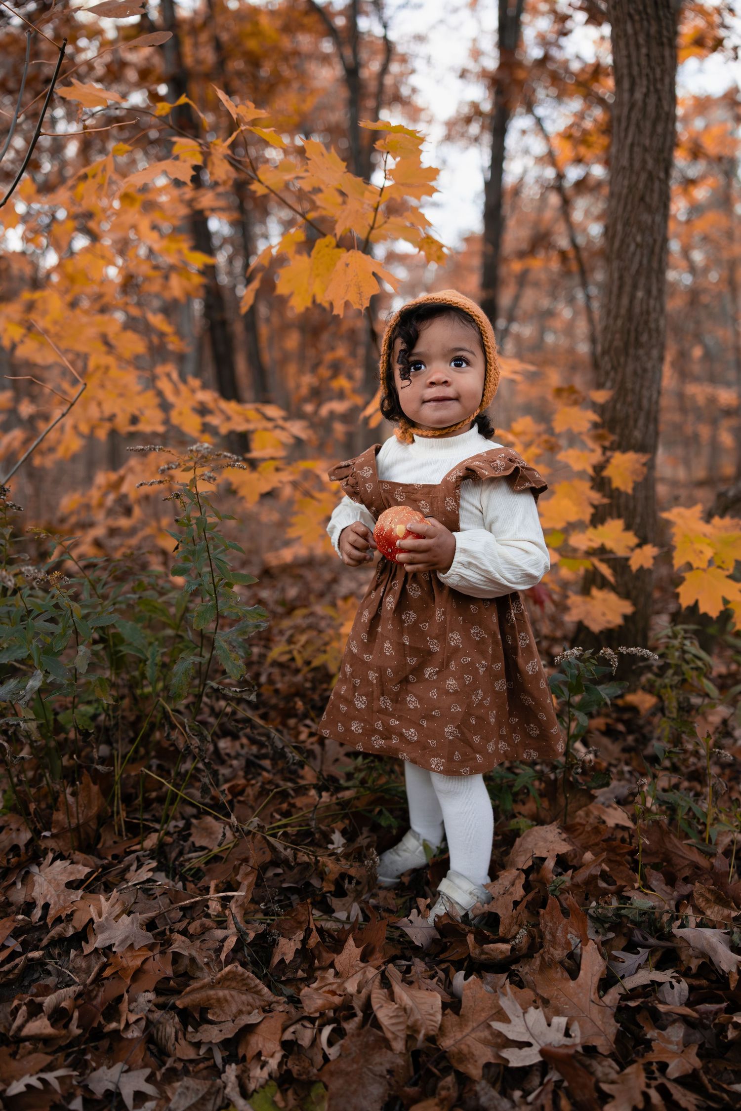 Toddler girl in brown floral dress and bonnet holding pumpkin among golden fall leaves.