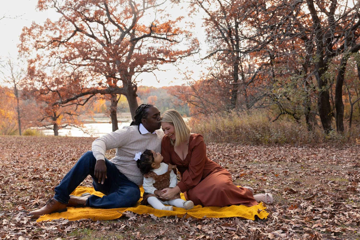 Happy interracial couple sitting on yellow blanket in autumn leaves with their dog by a lake.