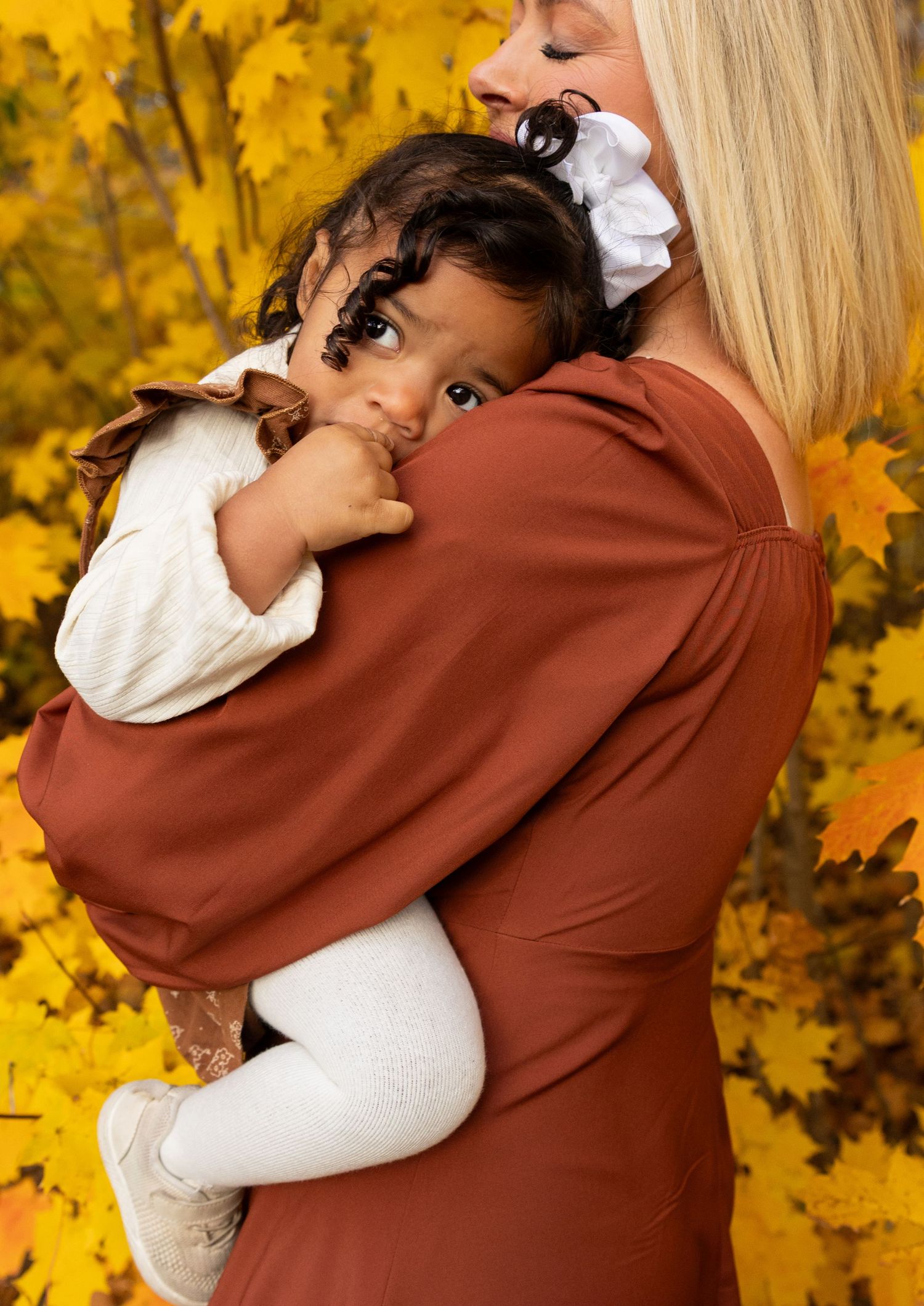 A blonde mother tenderly holds her curly-haired toddler against a vibrant yellow autumn leaves backdrop.