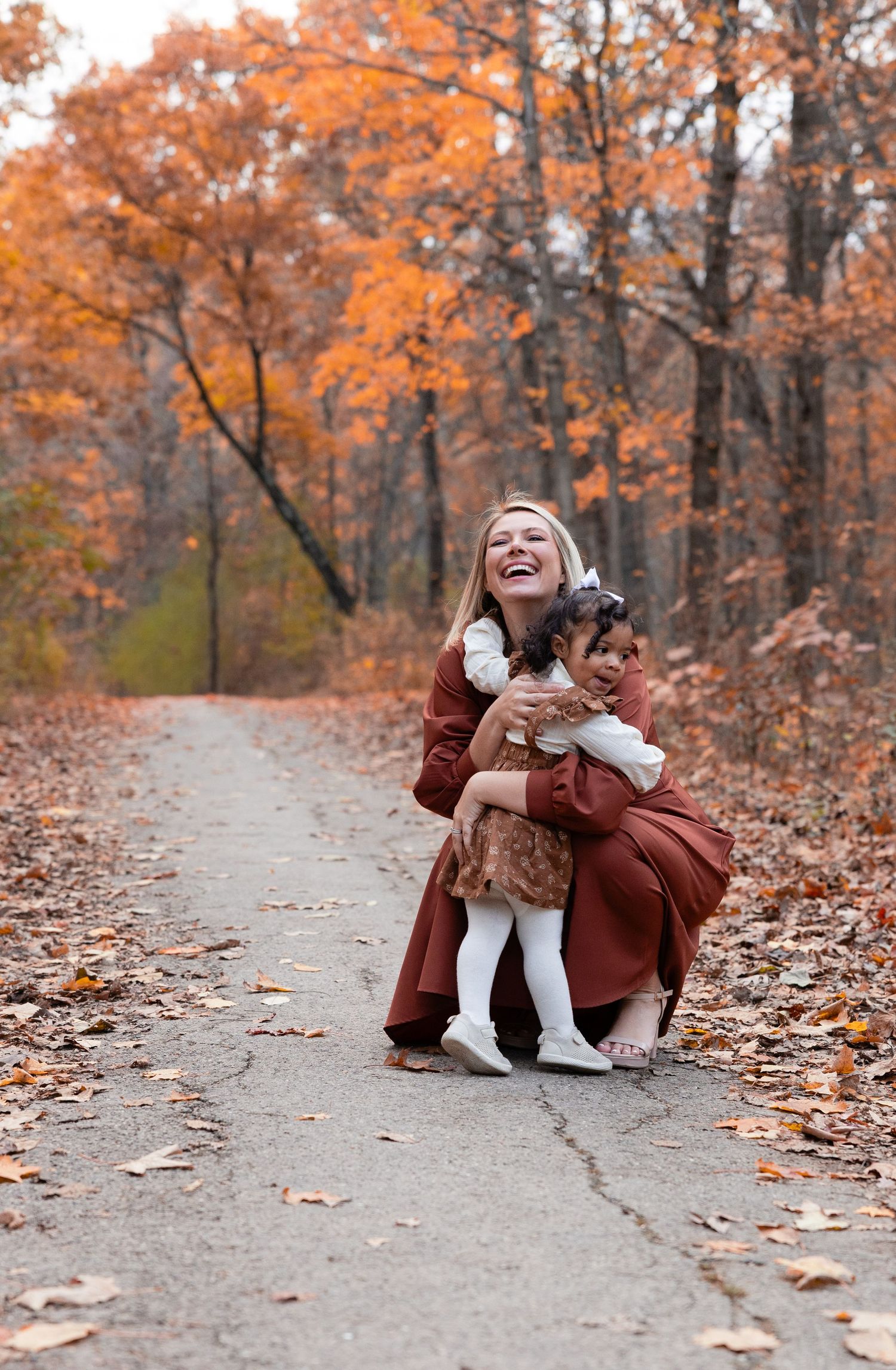 A laughing mother hugs her toddler daughter on a leaf-covered path surrounded by colorful fall foliage.