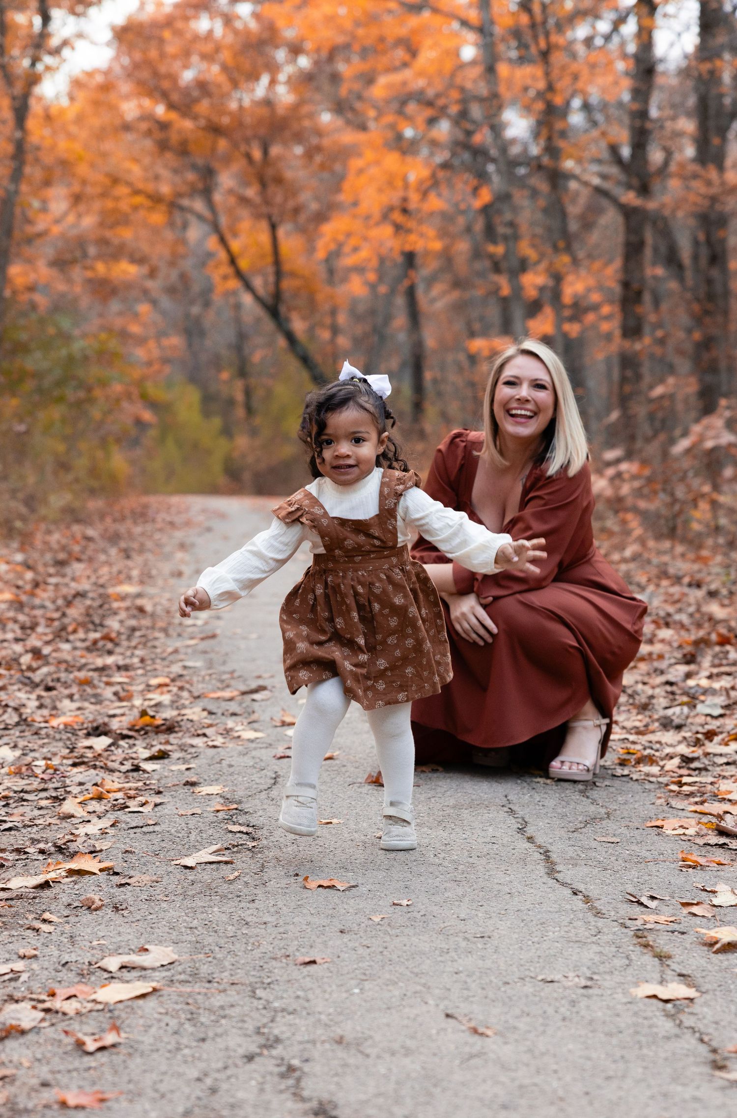 A blonde mother crouches beside her toddler daughter walking on an autumn forest path lined with fallen leaves.