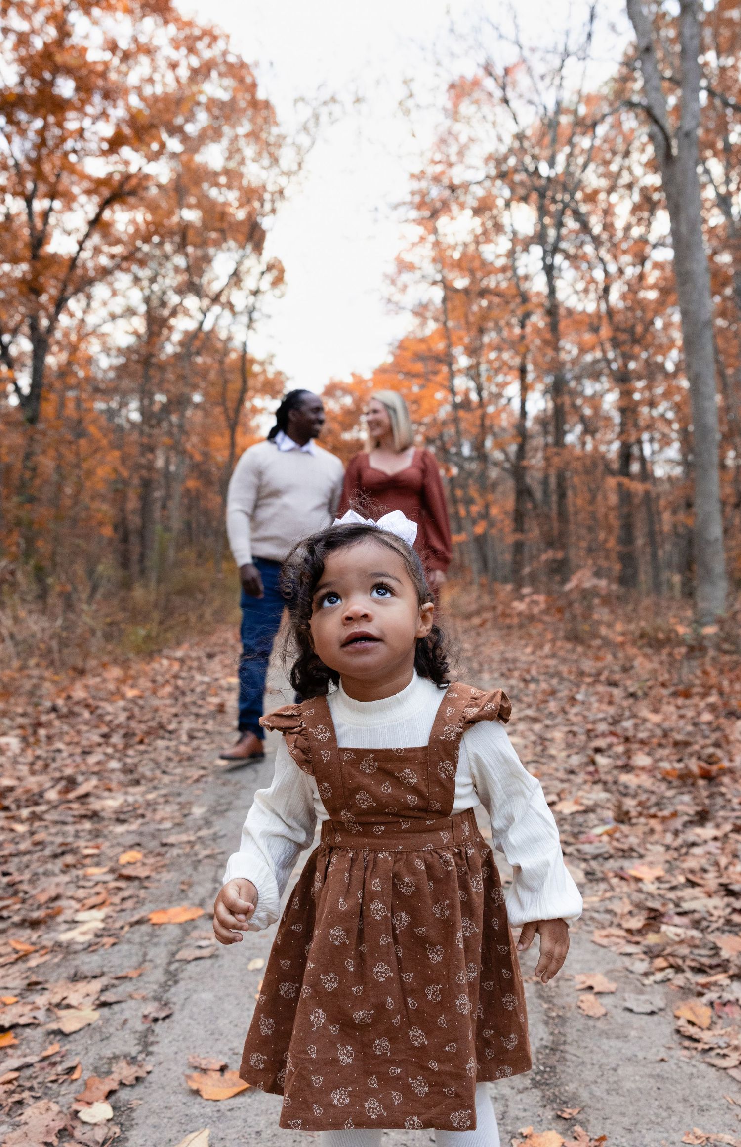 An adorable toddler girl in a brown dress explores a fall forest path while her parents stand behind her.
