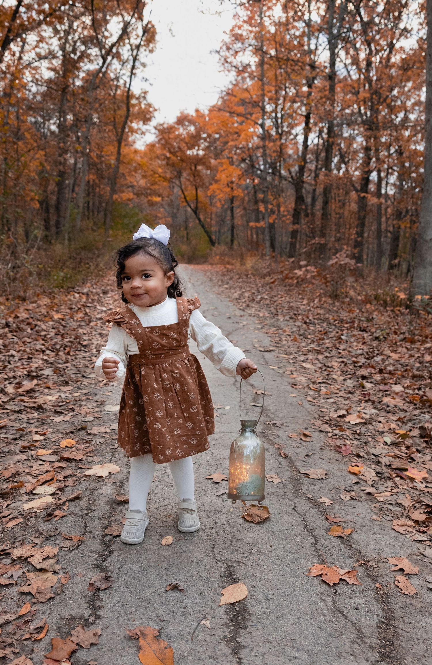 A cute toddler girl in a brown dress holds a glowing lantern on a leaf-covered autumn forest path.