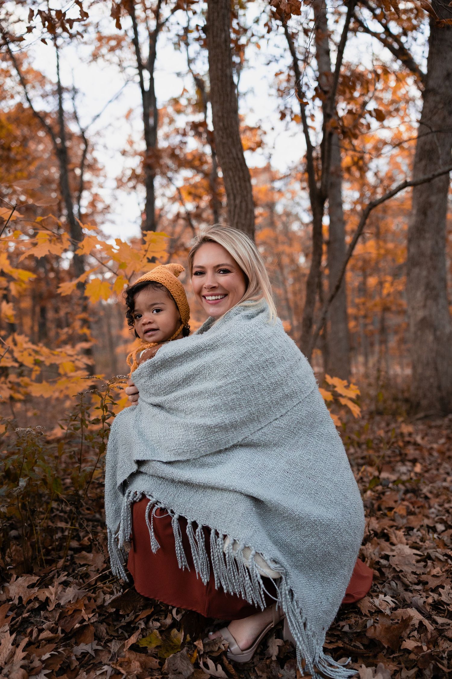Mother and toddler wrapped in gray blanket smiling in autumn forest.