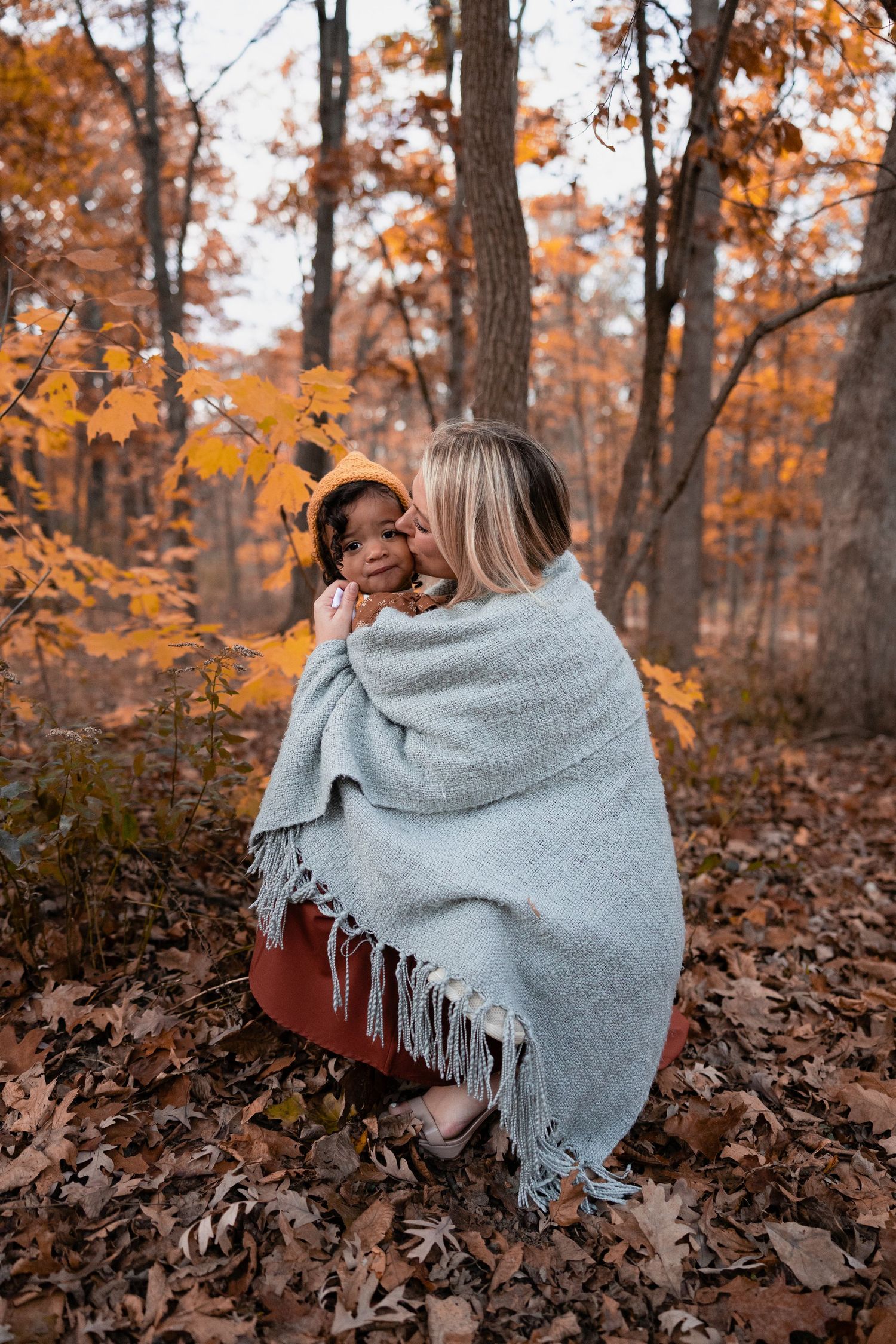 Mother kissing toddler wrapped together in gray blanket in autumn forest.
