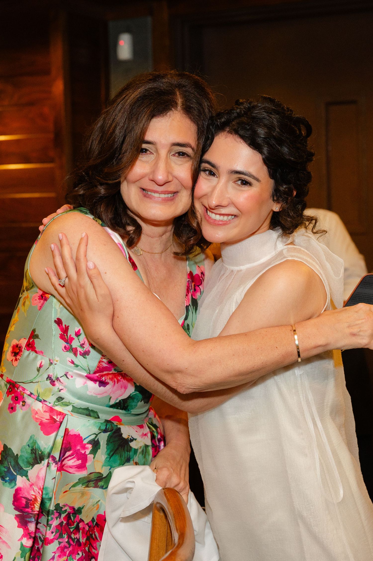 Two women smiling and hugging warmly, one in a floral dress and one in a white top, at an indoor event.