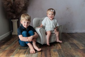 A toddler girl sits in a small gray chair beside her older brother on a wood floor in a studio portrait.