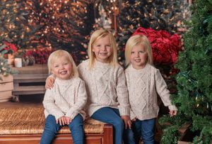 Three blonde sisters in matching cream sweaters posing by Christmas tree decorations.