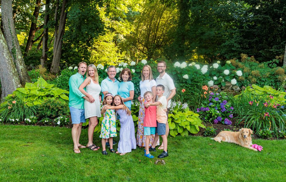 A large family of nine poses together in a lush green garden with colorful flowers and a golden retriever.
