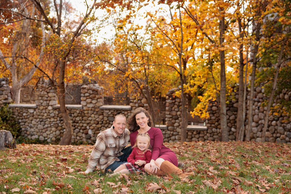 A family of three sits together on fallen autumn leaves in front of a stone wall outdoors.