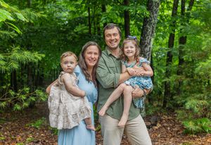 Family of four smiling outdoors in lush green forest, parents holding two young daughters in summer outfits.