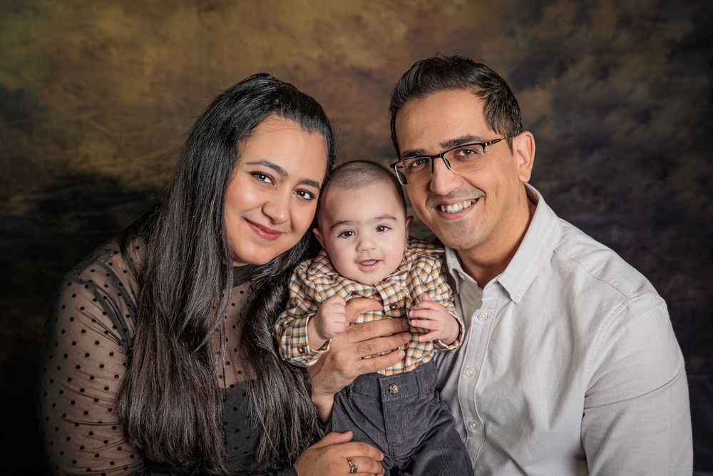 A smiling couple holds their happy baby in a professional studio portrait with a brown backdrop.