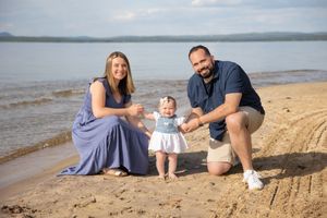 Young couple helping their baby girl take first steps on a sandy lakeside beach. Ellacoya State Park, Gilford NH.