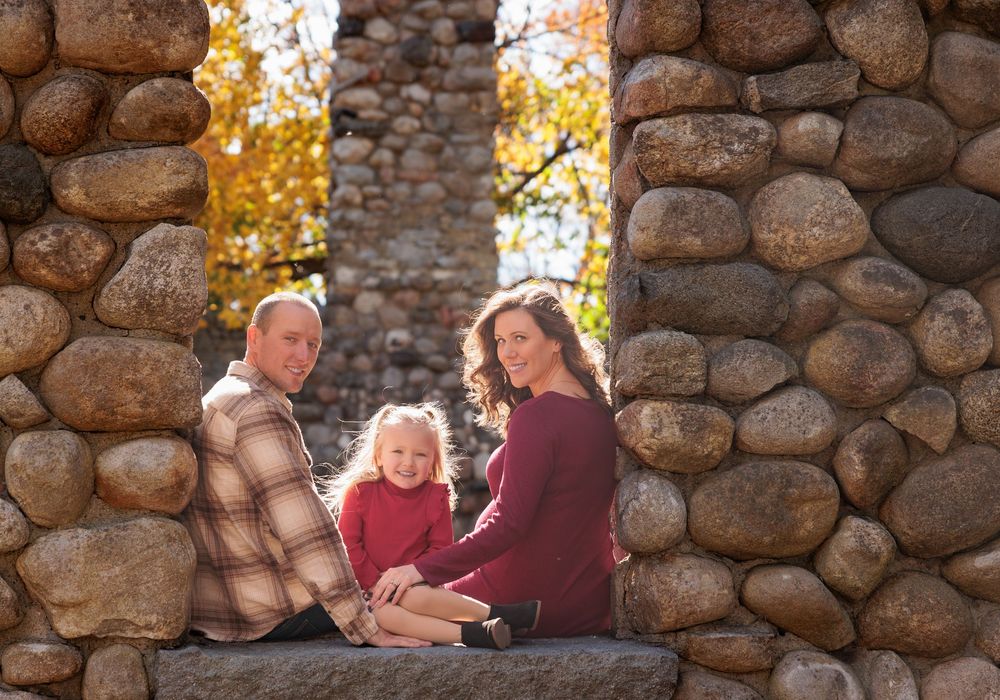 A family of three sits together framed by a rustic stone arch surrounded by autumn foliage.