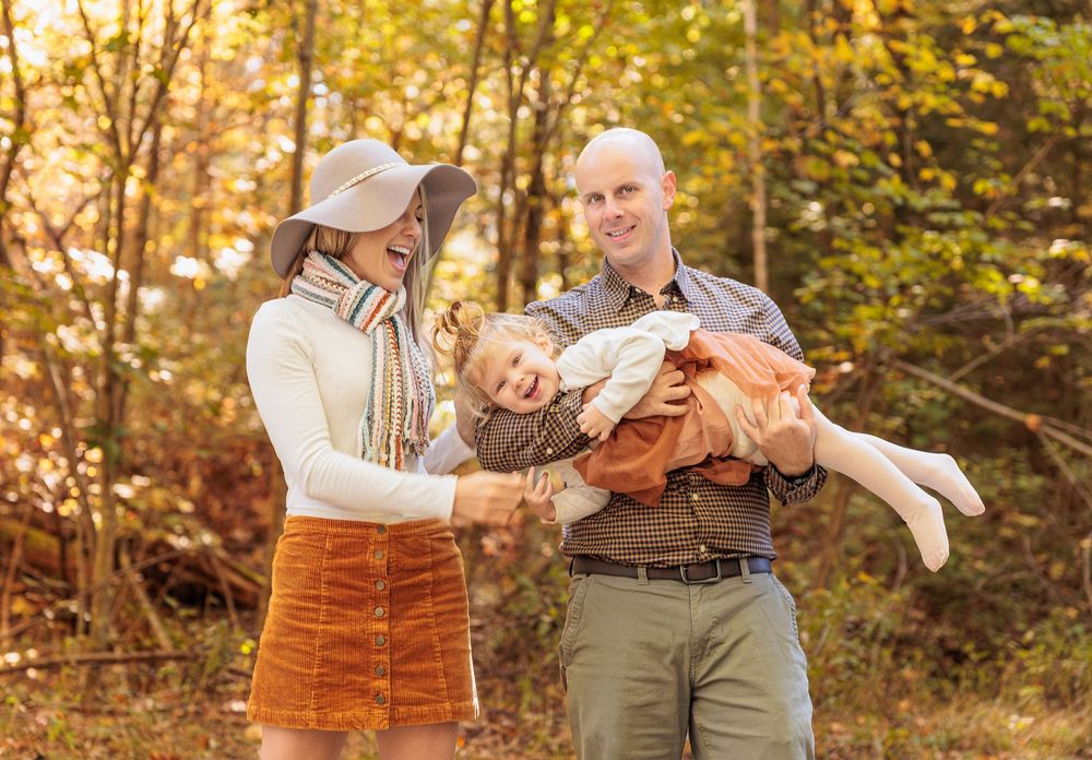 Parents laugh as they lift their young daughter in a sunny autumn forest family portrait. Northwood Meadows State Park, Northwood NH.