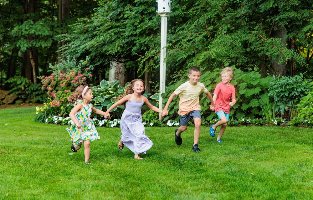 Four children hold hands and run joyfully across a lush green lawn in a sunny backyard.