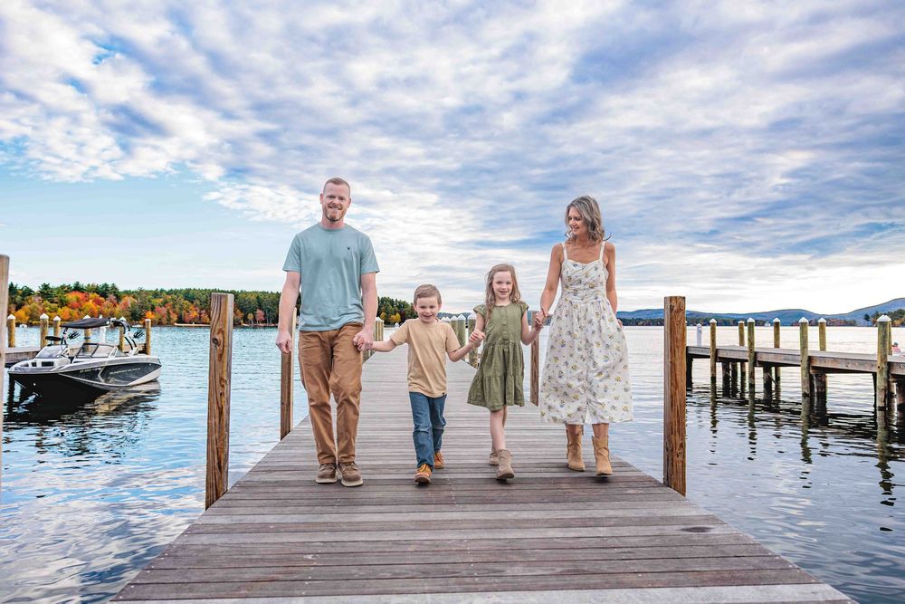 Happy family of four walking hand-in-hand on a lakeside dock under a beautiful cloudy sky. Cate Park Wolfeboro NH