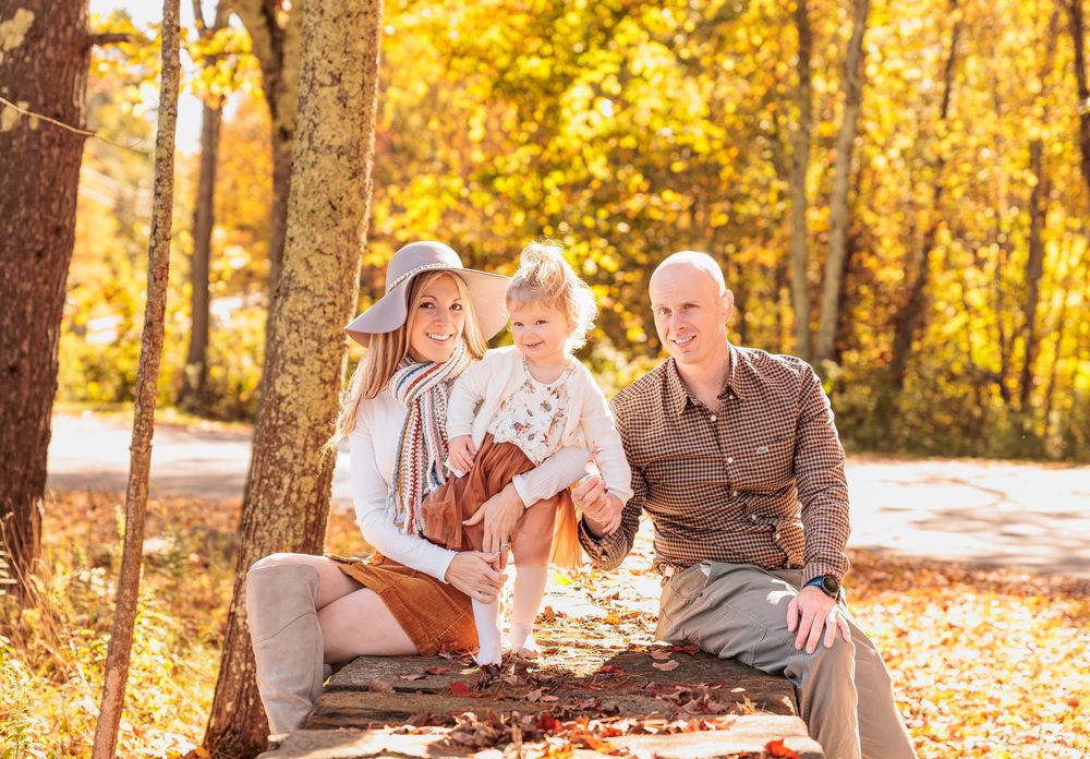 A family of three sits on a bench beneath a tree surrounded by vibrant golden fall foliage. Northwood Meadows State Park, Northwood NH.