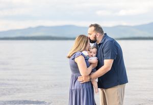 Couple sharing kiss over baby they hold together at lakeside with mountains behind. Ellacoya State Park, Gilford NH.