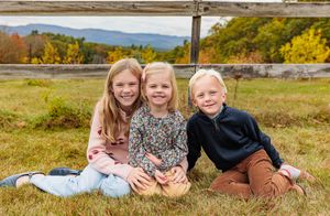 Three young siblings sitting together in grassy field with autumn mountains behind.