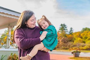 Mother holding laughing toddler girl in teal dress outdoors in autumn setting. Alton Bay NH.