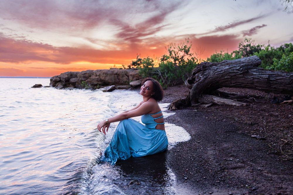 Woman in blue dress sitting at water's edge during vibrant sunset with dramatic orange sky and rocky shoreline.