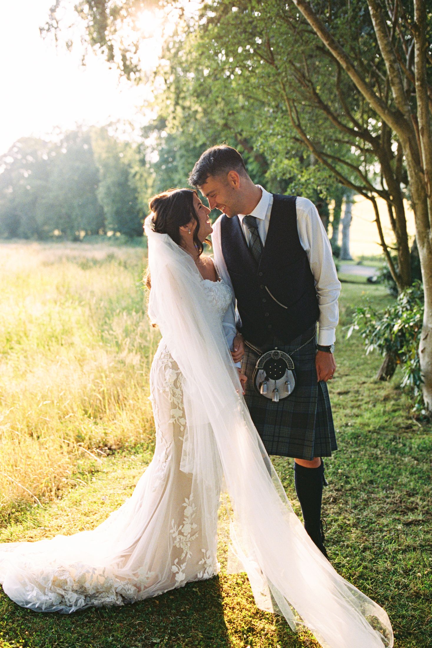 Bride in lace gown and kilted groom share a romantic moment in golden sunlight.