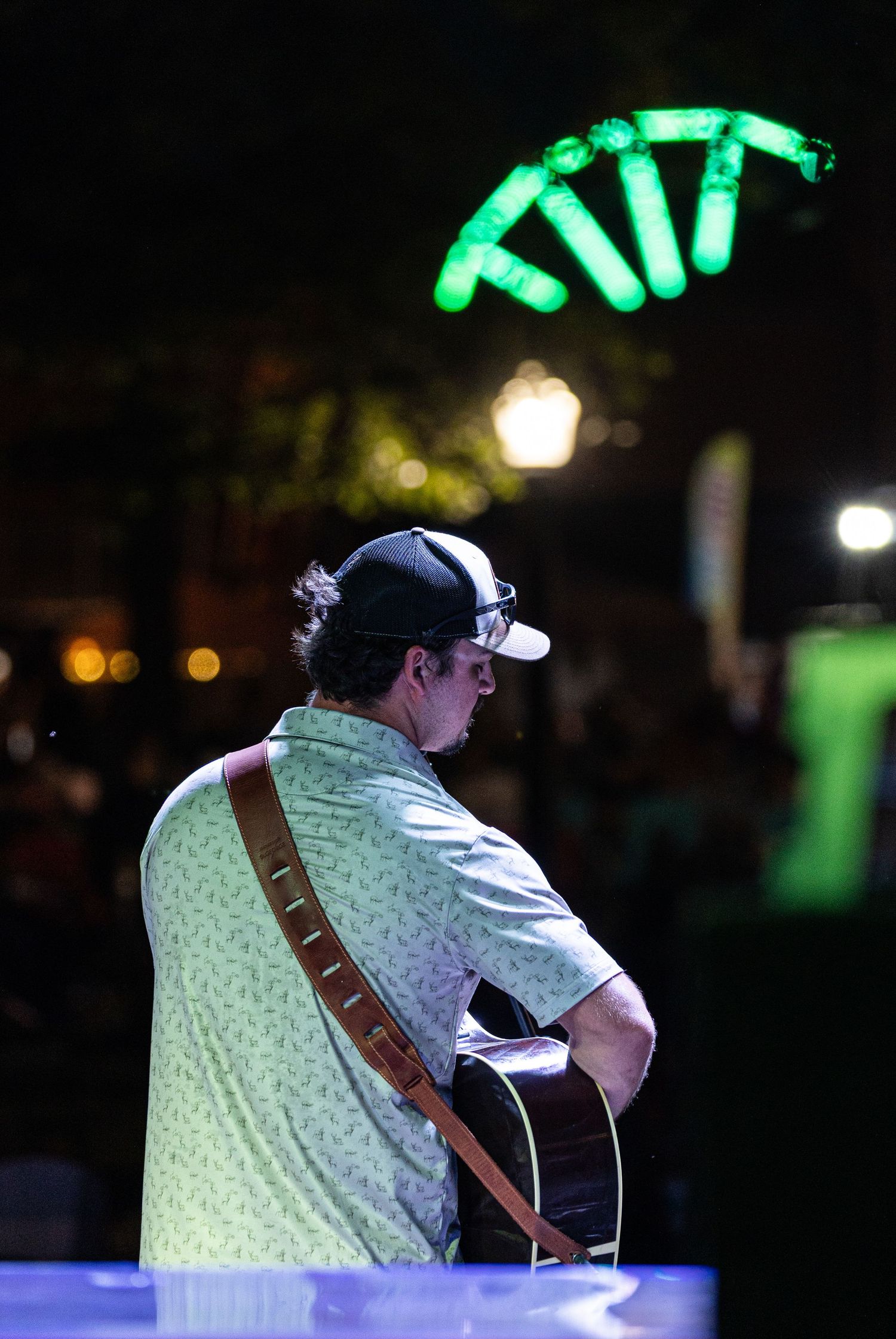 Musician playing acoustic guitar at an outdoor night event with green neon lights glowing in the background.