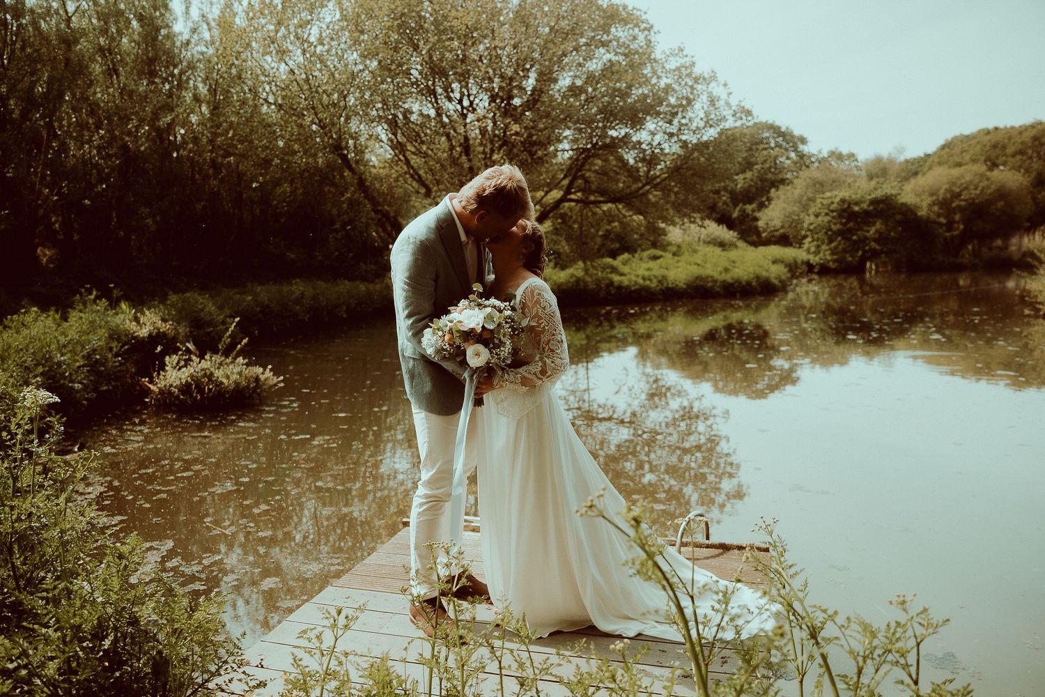 Bride and groom sharing a romantic kiss by a serene pond on their wedding day.