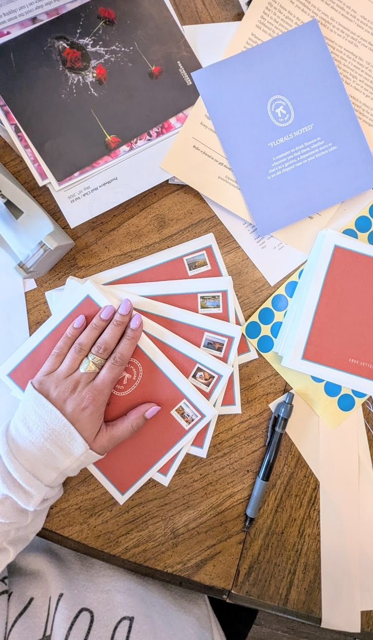 A hand with rings holds red envelopes with stamps on a wooden desk surrounded by papers, blue dots, and a pen.