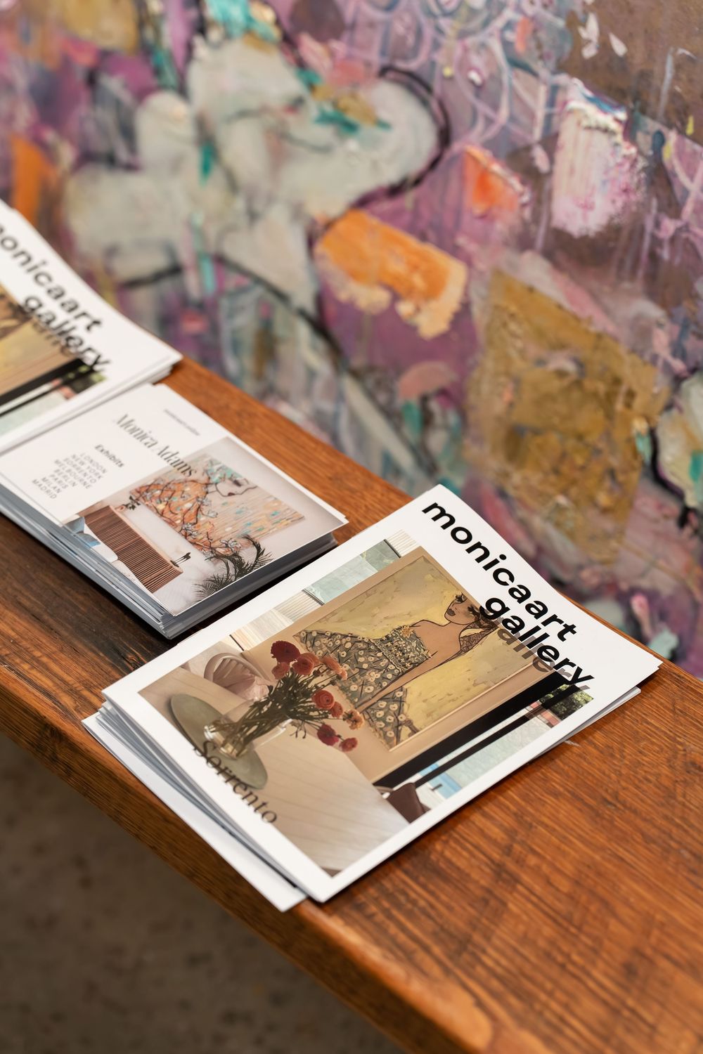 Monicaart Gallery brochures displayed on a wooden table against a colorful abstract painted wall backdrop.