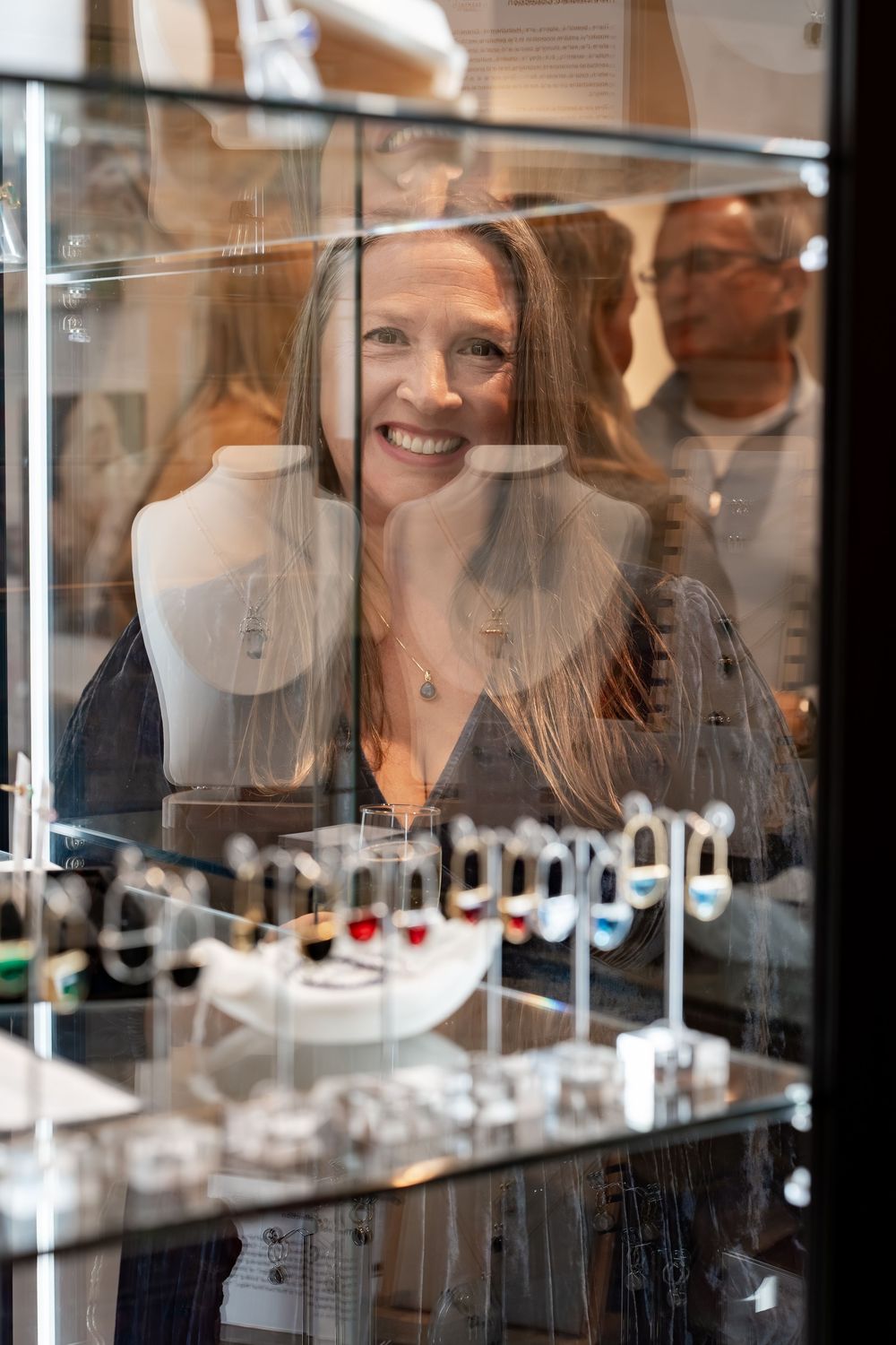 Smiling woman browsing jewelry display case filled with rings and necklaces at an art gallery event.