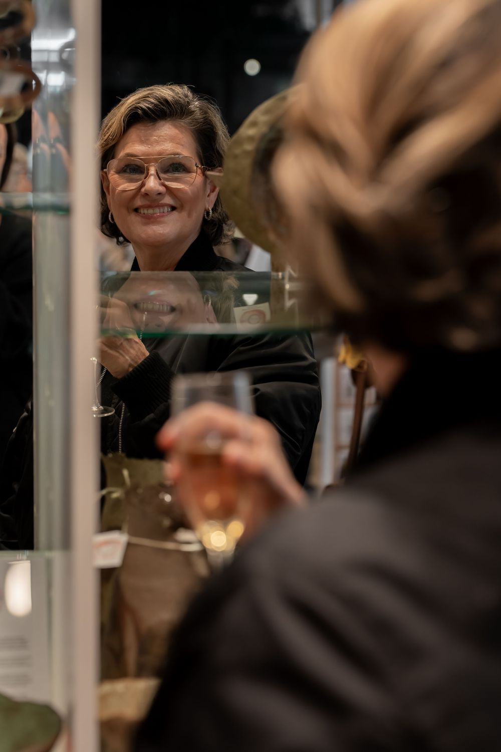 Two women socializing at an event, one smiling warmly while the other holds a glass of white wine.