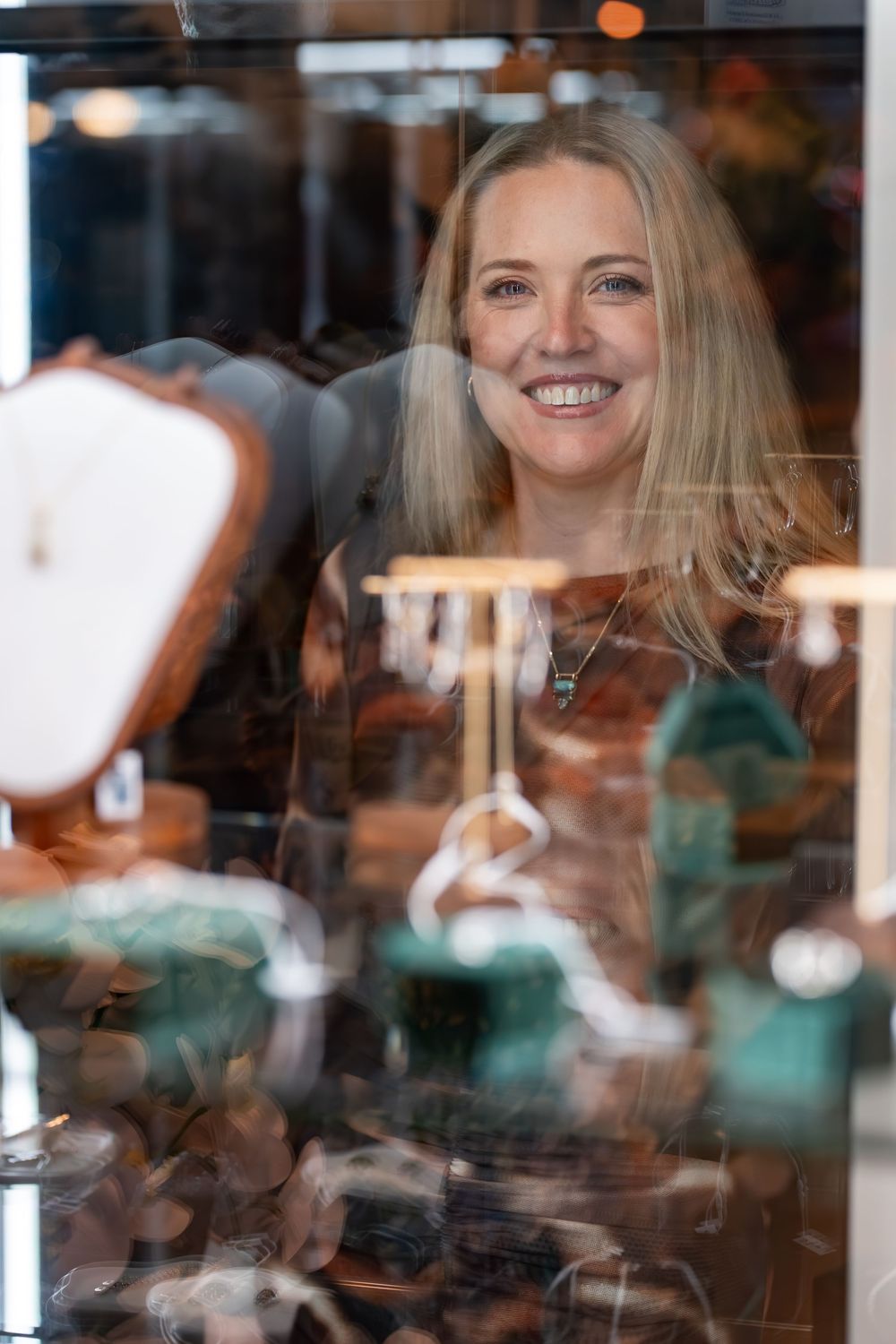 Smiling blonde woman viewed through a jewelry display case filled with gemstone pieces and elegant accessories in a boutique.