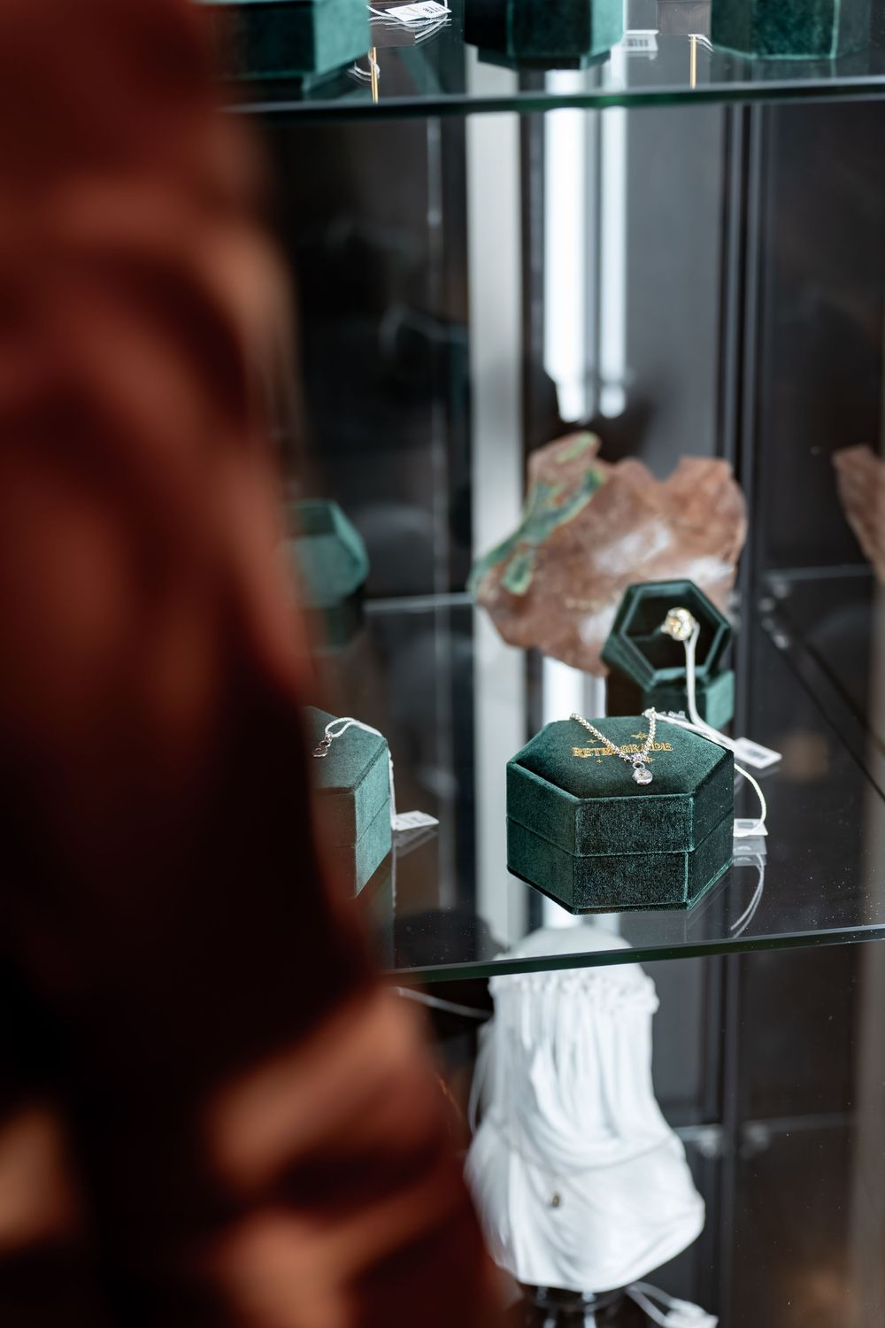 Person browsing a jewelry display case with green velvet ring boxes and decorative items on glass shelves.