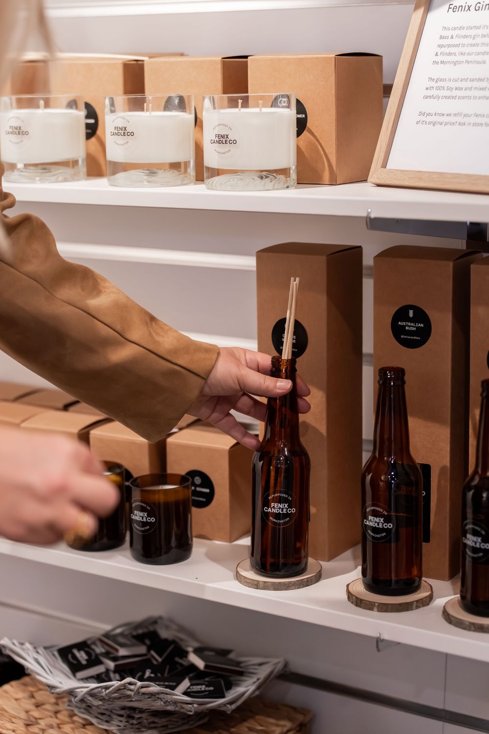 Person arranging amber bottle reed diffusers and candles on white retail shelves in a modern gift shop.