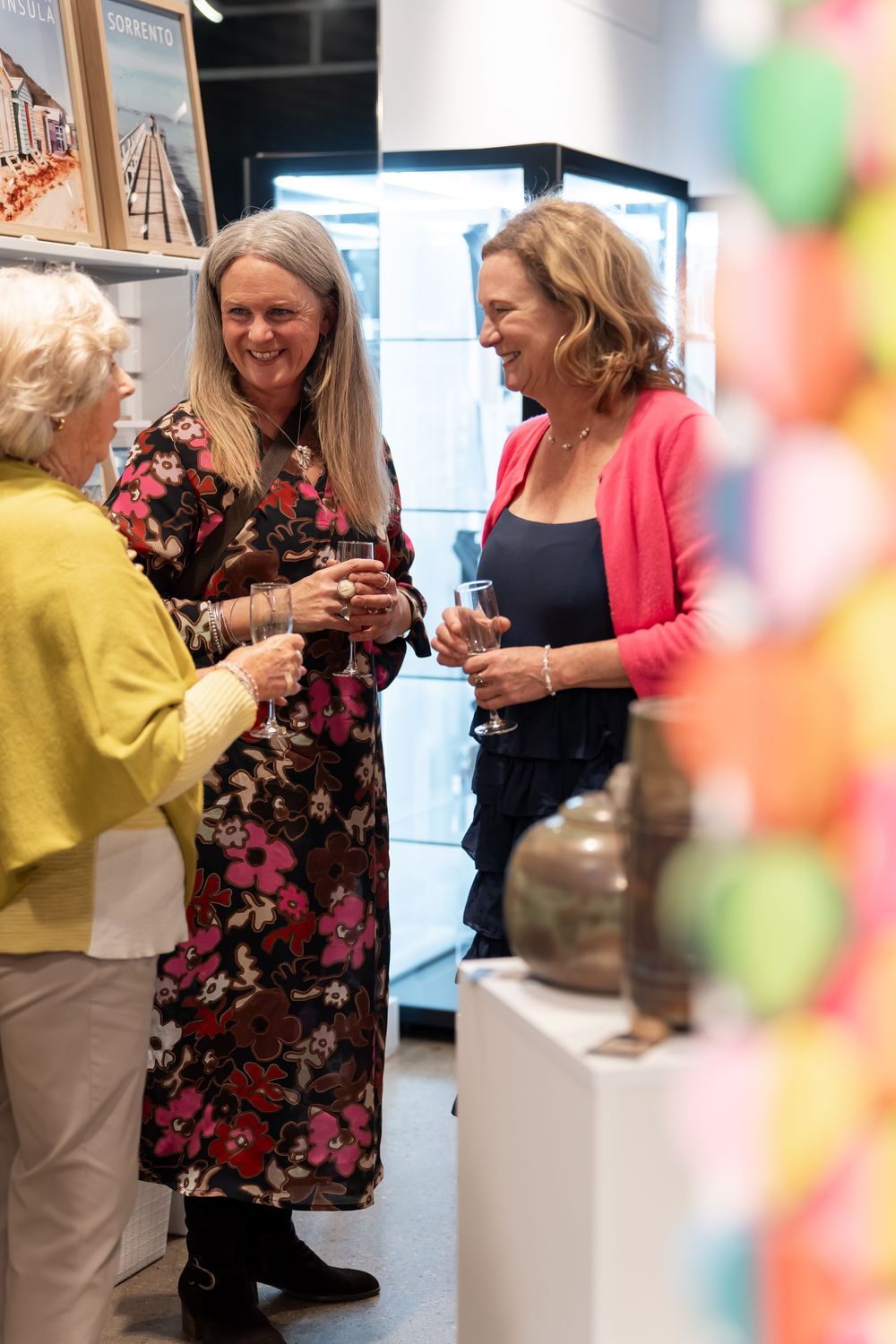 Three women socializing at an art gallery event, holding wine glasses and smiling together.