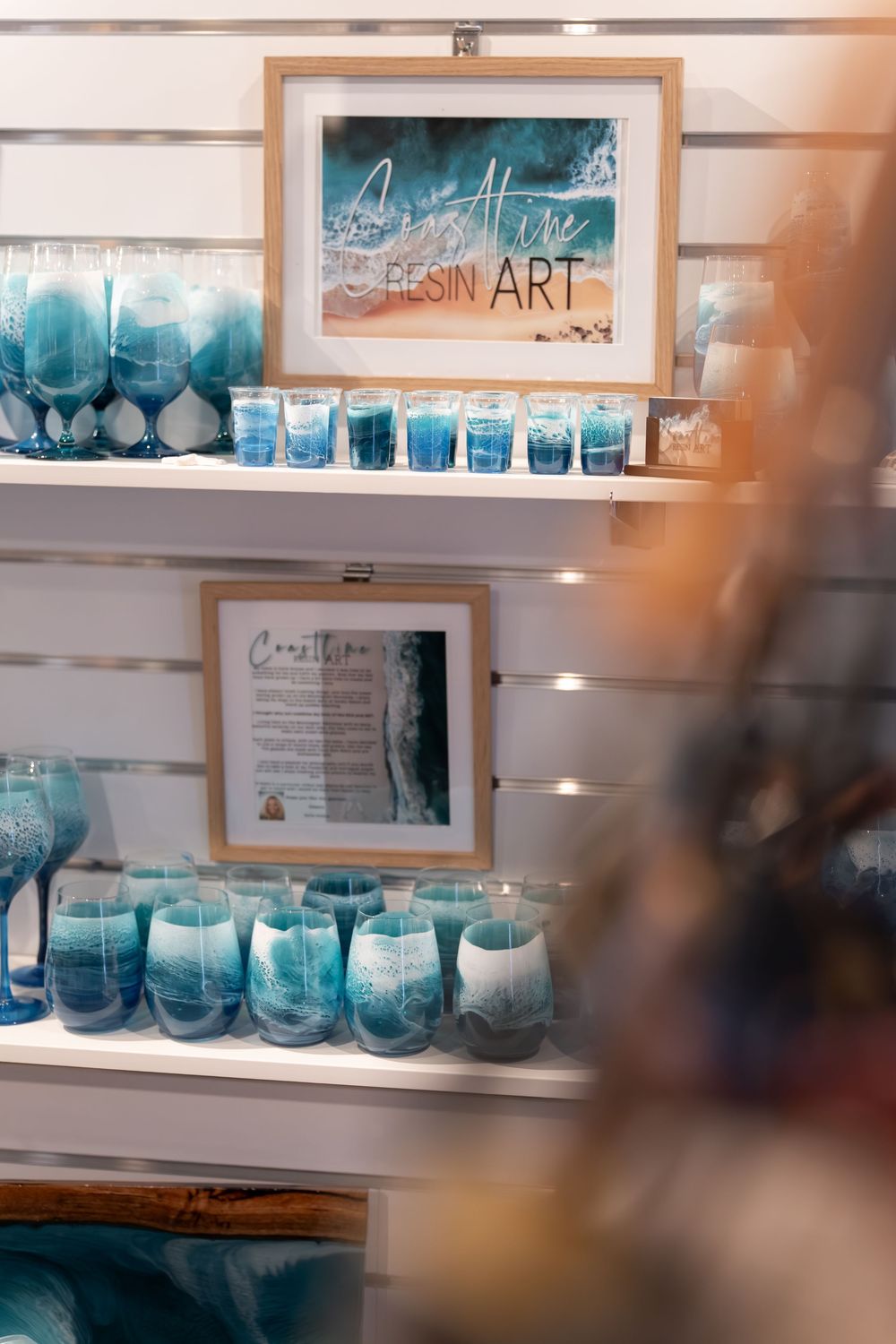 Coastal resin art shop display with blue ocean-themed glassware and framed artwork on white shelves.