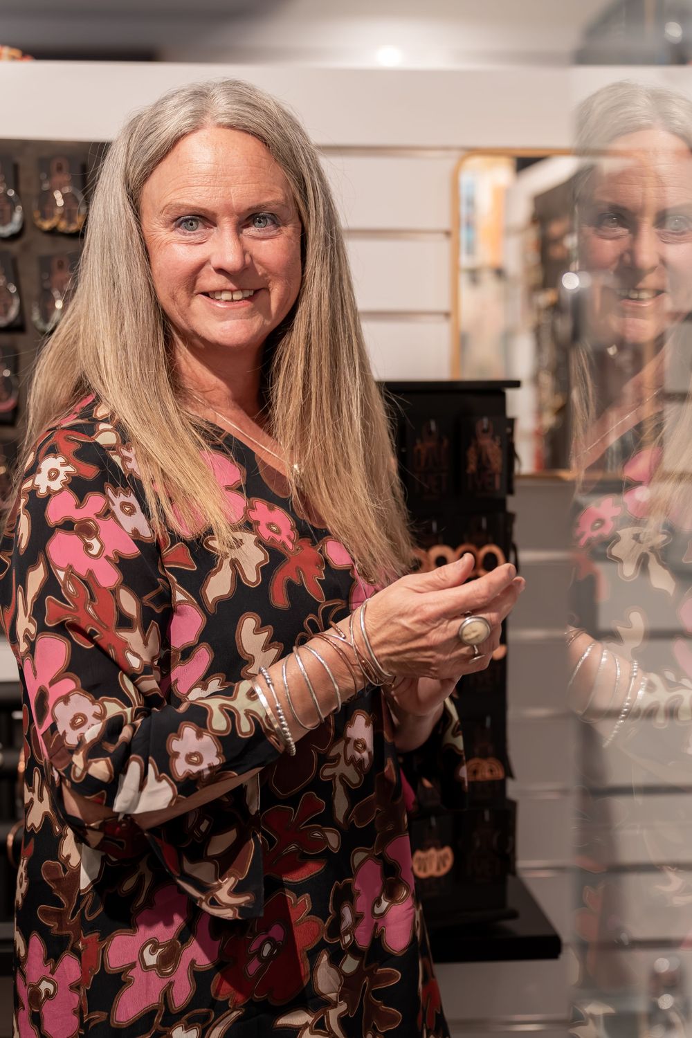 Smiling woman with long gray hair in floral dress holding jewelry in a retail store.
