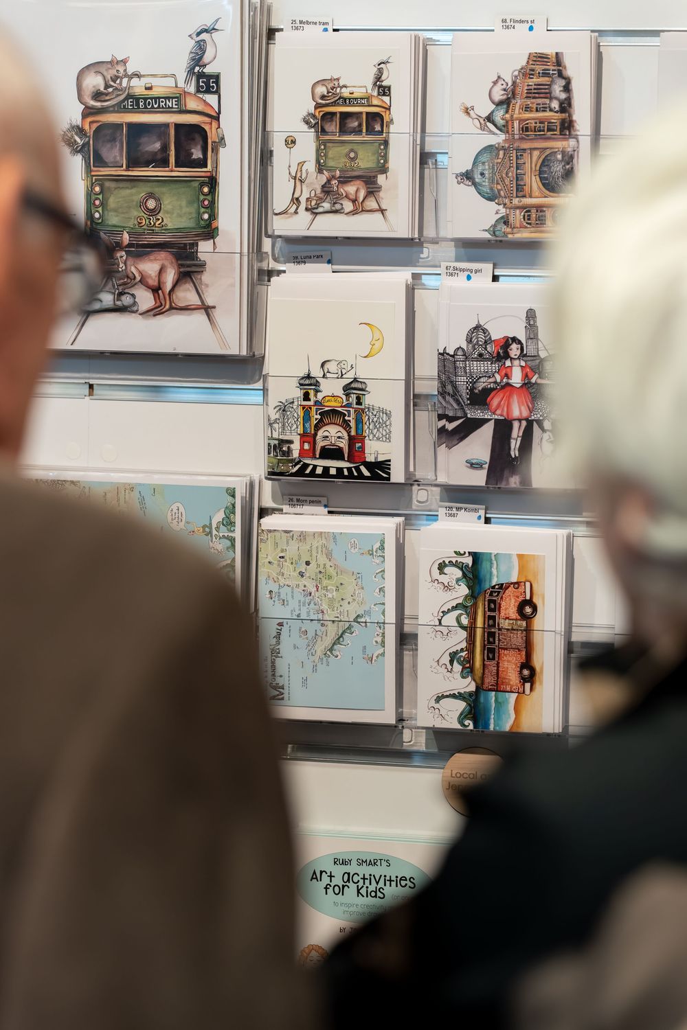 Two people viewing a display of illustrated art cards featuring whimsical trams, maps, and characters at an art exhibition.