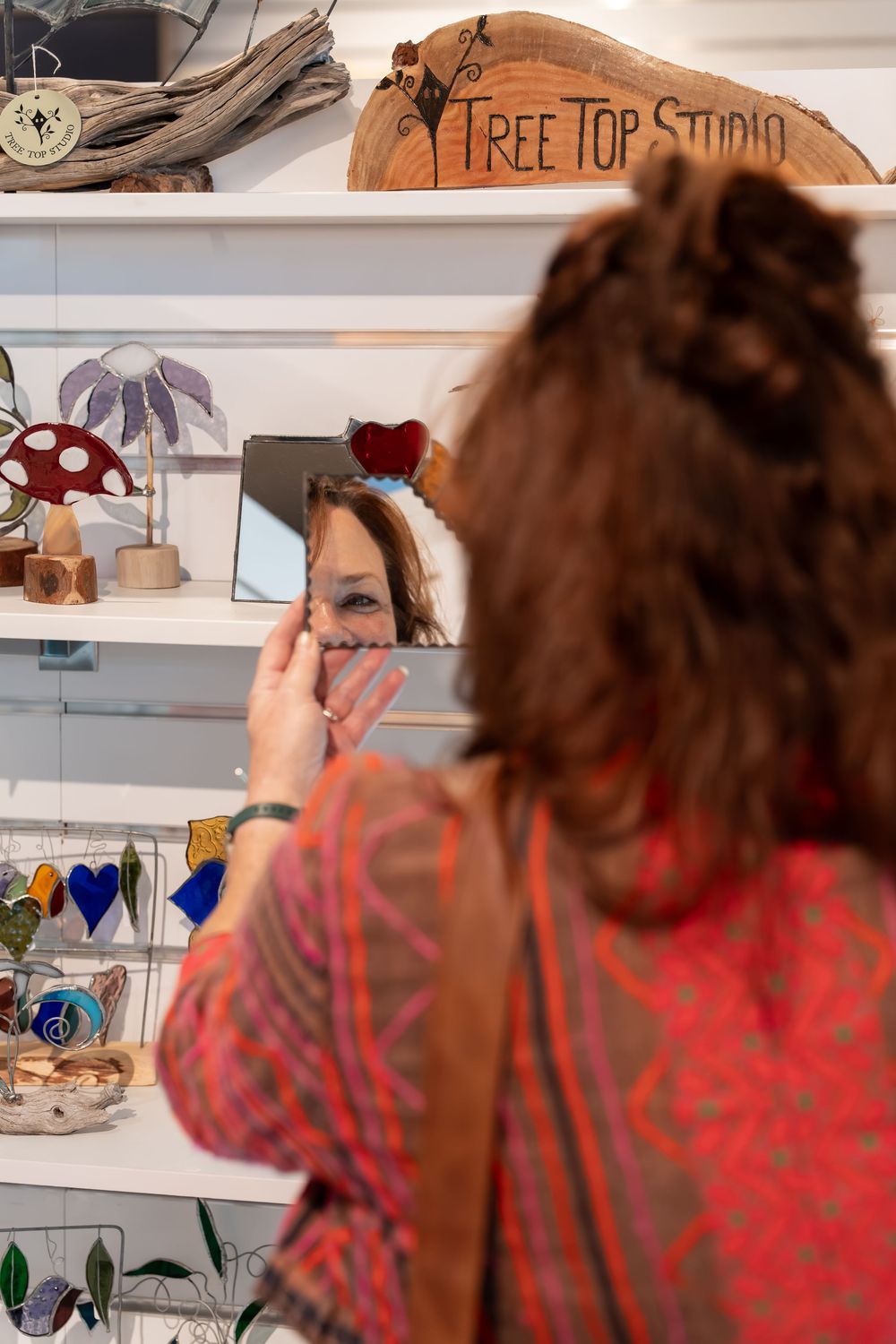 Woman trying on jewelry while looking in a mirror at Tree Top Studio craft shop display.