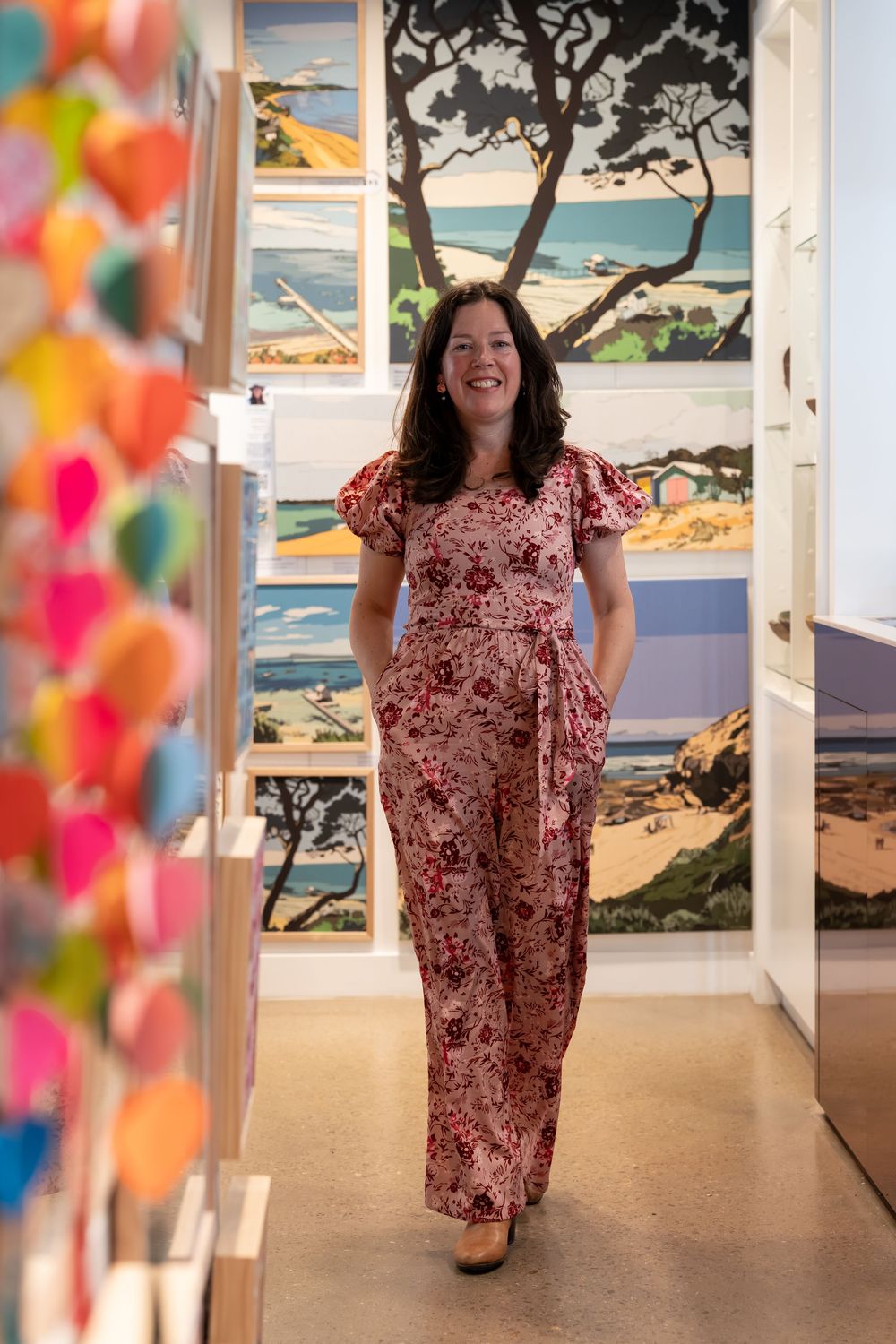 Woman in floral jumpsuit smiling in art gallery with colorful coastal landscape paintings on display.