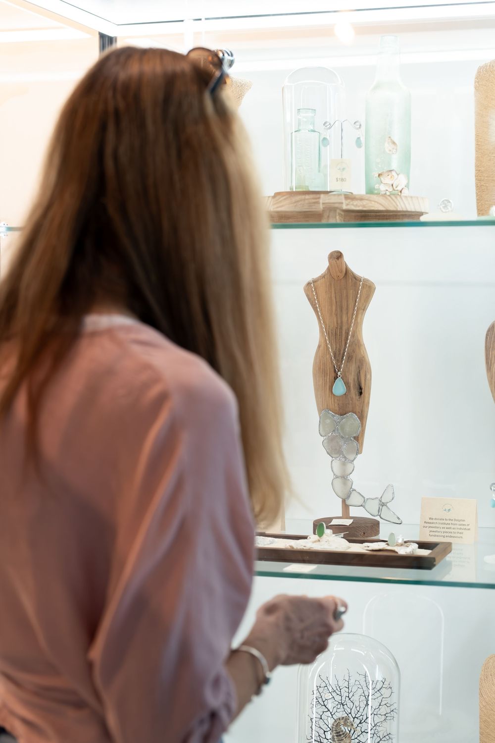 Woman browsing jewelry display case with turquoise necklace on mannequin bust in boutique shop.
