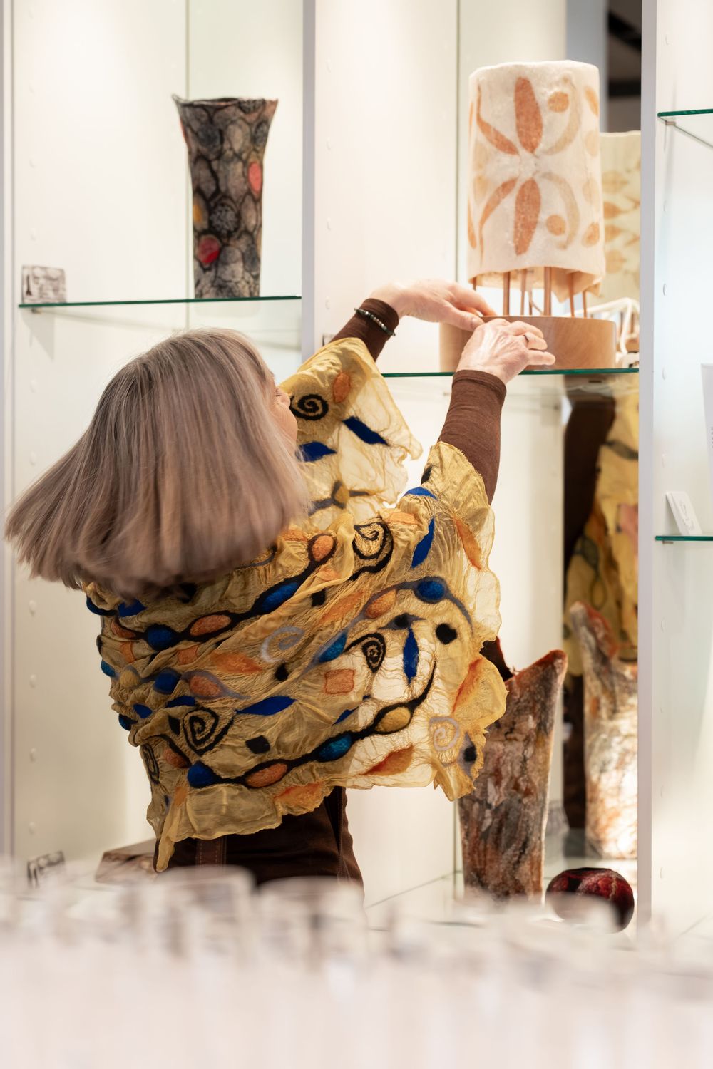 Woman in colorful patterned jacket arranging decorative vases on glass shelves in an art gallery.