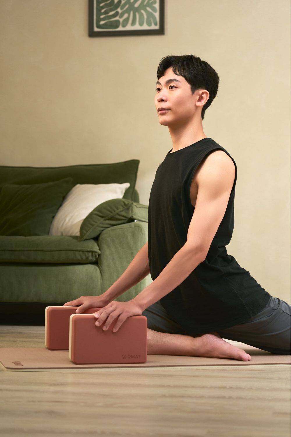 Man practicing yoga splits pose with blocks on mat in living room, wearing black sleeveless top.