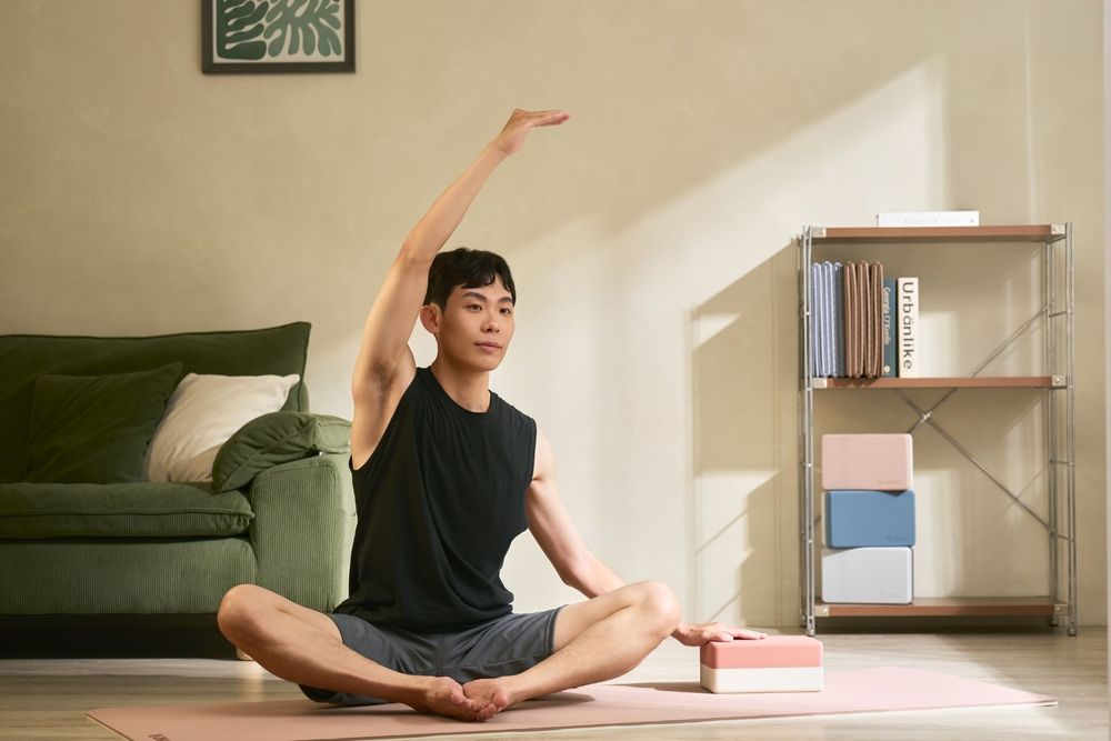 Young Asian man practicing yoga seated pose with arm raised on pink mat in modern living room.
